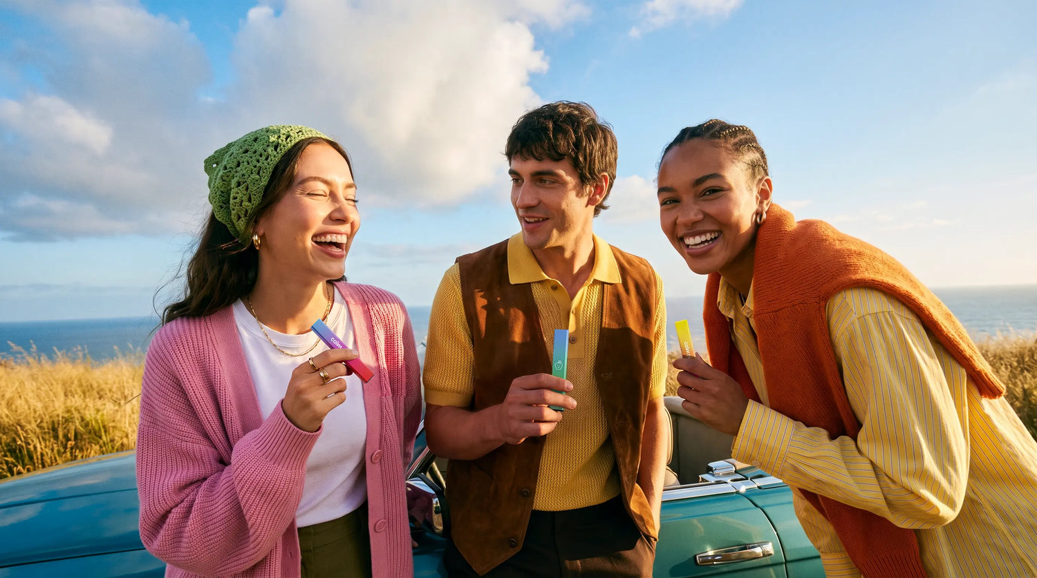 Three young adults laughing outdoors by a convertible car, each holding a brightly colored Calm Puffs personal aroma diffuser. They are dressed in colorful casual summer clothing, with a blue sky and ocean in the background.